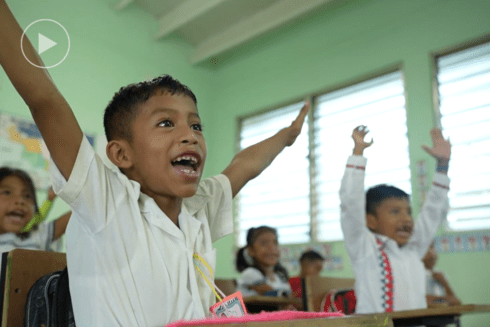 Two smiling children raise their hands while sitting at their desk at school - Investor - Inter American Development Bank - IDB
