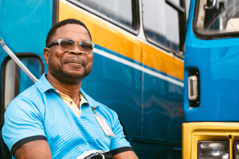 Smiling man wearing glasses and a striped shirt standing confidently in front of a colorful public bus - Inter-American Development Bank - IDB